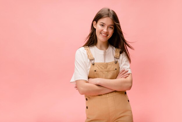 Sorridente jovem morena europeia com braços cruzados em camiseta e macacão em fundo rosa Conceito de estilo de vida