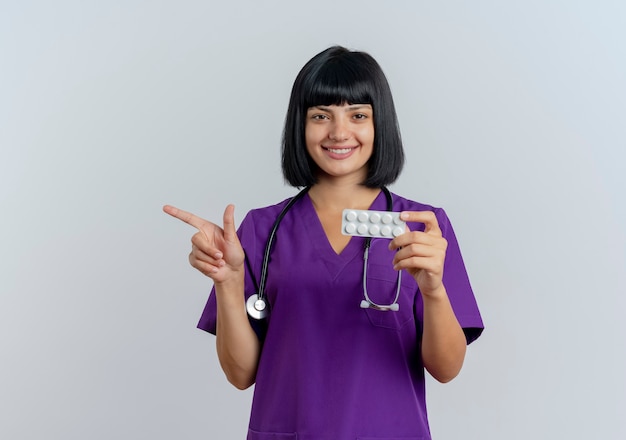 Sorridente, jovem morena médica de uniforme com estetoscópio segurando o remédio e apontando para o lado