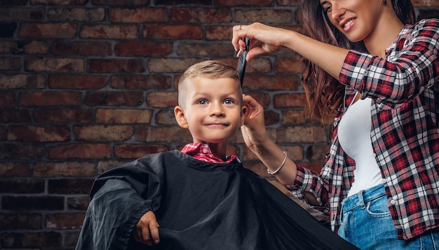 Sorridente menino pré-escolar cortando o cabelo. cabeleireiro de crianças com tesoura e pente está cortando garotinho na sala com interior loft.