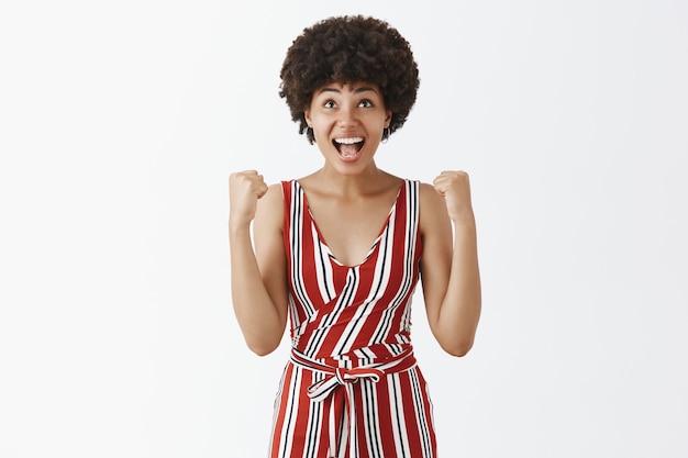 Sorridente modelo feminino atraente de pele escura em traje estiloso, parecendo animado, levantando os punhos em sucesso ou triunfo