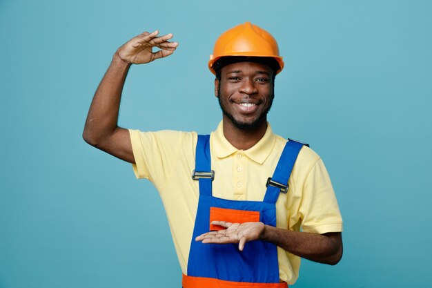 Sorridente mostrando tamanho jovem construtor afro-americano em uniforme isolado em fundo azul