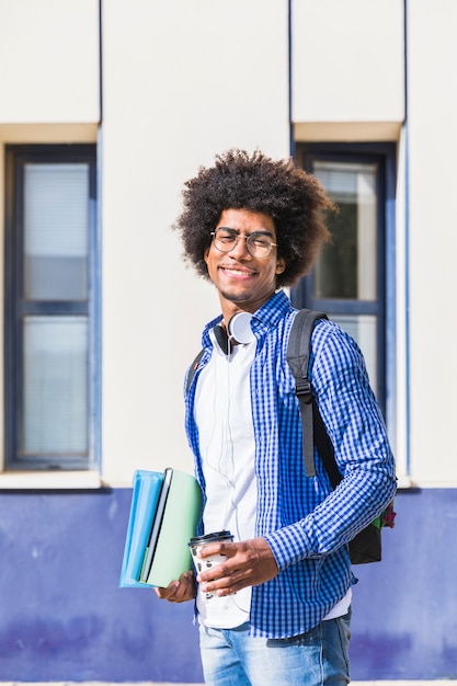 Sorrindo, adolescente, estudante masculino, carregar, mochila, ligado, ombro, segurando, pilha, de, livro, e, takeaway, xícara café, ficar, em, campus