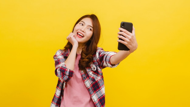 Sorrindo adorável fêmea asiática fazendo selfie foto no smartphone com expressão positiva em roupas casuais e olhando para a câmera sobre parede amarela. Mulher feliz adorável feliz alegra sucesso.