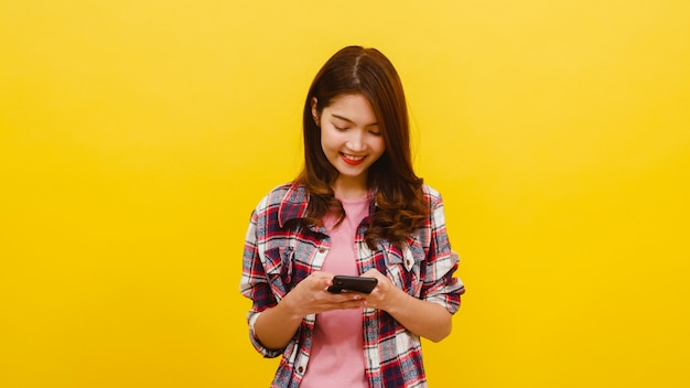 Sorrindo adorável fêmea asiática usando telefone com expressão positiva, sorri amplamente, vestido com roupas casuais e olhando para a câmera sobre parede amarela. Mulher feliz adorável feliz alegra sucesso.