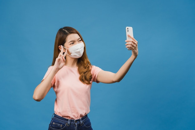 Sorrindo adorável mulher asiática usando máscara médica fazendo selfie foto no telefone inteligente com expressão positiva em roupas casuais e suporte isolado na parede azul