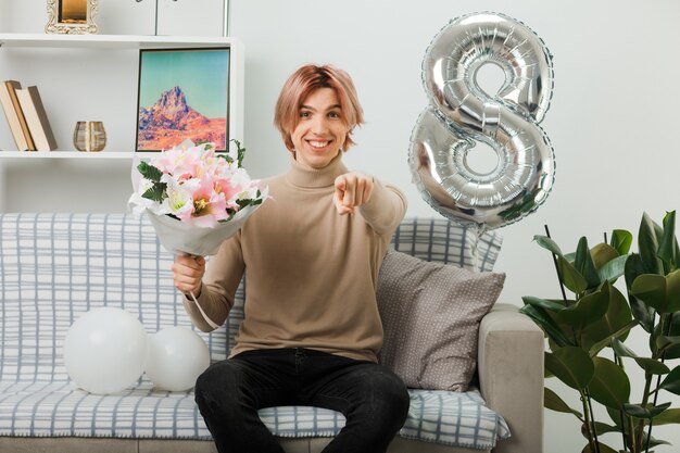 Sorrindo, apontando para um cara bonito no dia da mulher feliz, segurando um buquê sentado no sofá da sala de estar