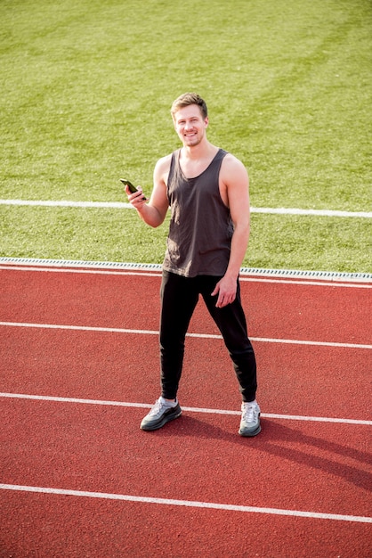 Sorrindo atleta do sexo masculino em pé na pista de corrida, segurando o telefone celular na mão
