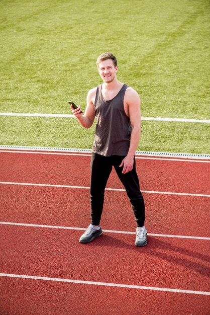 Sorrindo atleta do sexo masculino em pé na pista de corrida, segurando o telefone celular na mão