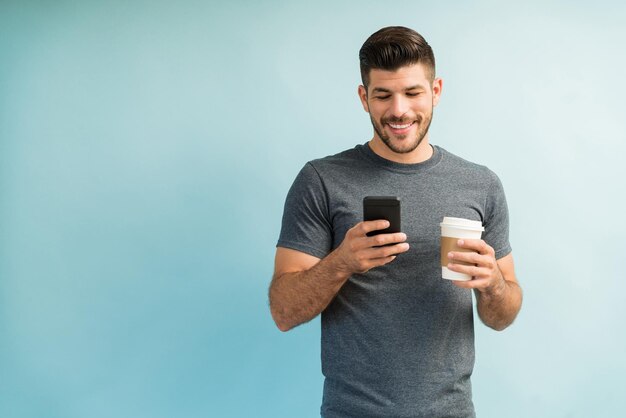 Sorrindo atraente homem latino enviando mensagens de texto no smartphone e segurando a xícara de café em pé contra um fundo liso