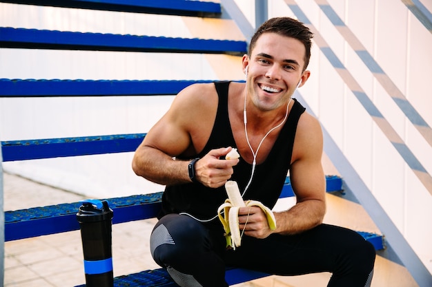 Foto grátis sorrindo cara esportiva em fones de ouvido, comendo uma banana, depois de treino, sentado na escada