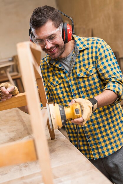Sorrindo carpinteiro masculino usando lixadeira elétrica para madeira na bancada