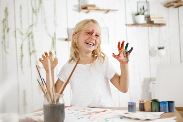 Sorrindo com os dentes loirinha, mostrando as mãos na pintura. Menina alegre de sete anos de idade, ocupada com um desenho de tinta sem bagunça.