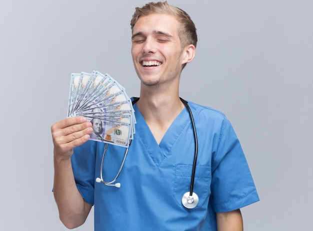 Sorrindo com os olhos fechados, jovem médico vestindo uniforme de médico com estetoscópio segurando dinheiro isolado na parede branca