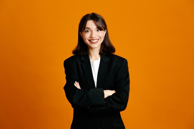 Sorrindo cruzando as mãos jovem linda fêmea vestindo jaqueta preta isolada em fundo laranja