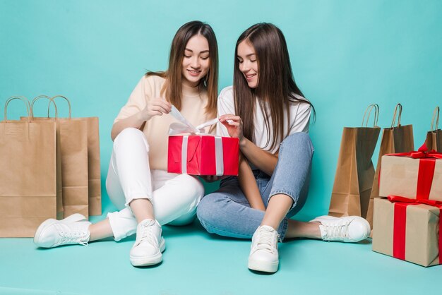 Sorrindo, duas meninas sentadas sacolas de compras no chão e presentes abertos na parede turquesa.