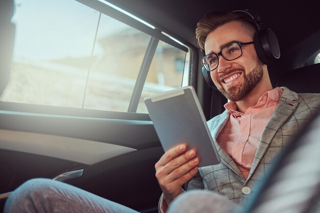 Sorrindo elegante jovem empresário de terno cinza e camisa rosa, ouvindo música em fones de ouvido enquanto estiver usando um computador tablet, andando no banco de trás em um carro de luxo.