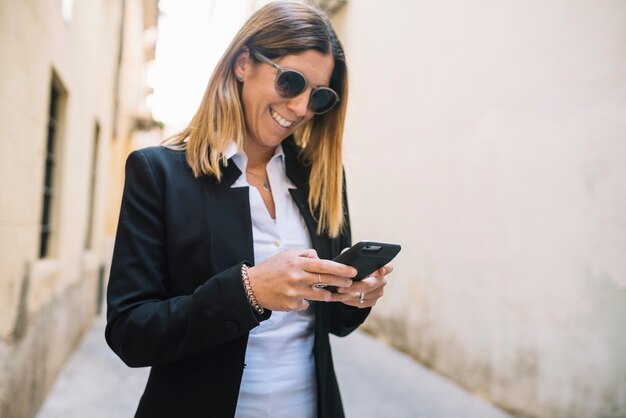 Sorrindo, elegante, mulher jovem, usando, smartphone, entre, edifícios, ligado, rua