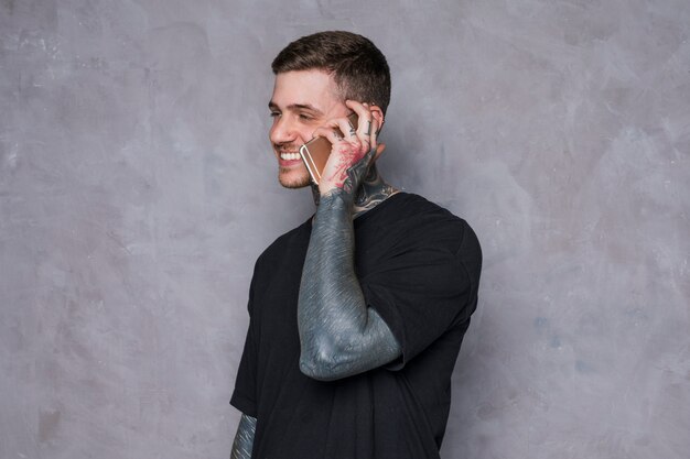 Sorrindo, homem jovem, com, tatuagem, ligado, seu, mão falando, ligado, telefone móvel, contra, parede cinza