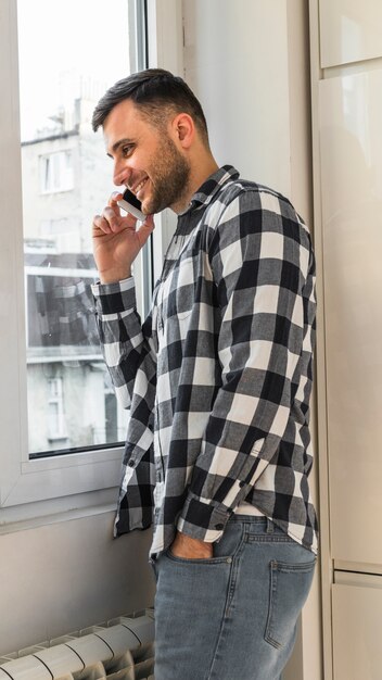 Sorrindo, homem jovem, falando telefone móvel, ficar, perto, a, janela