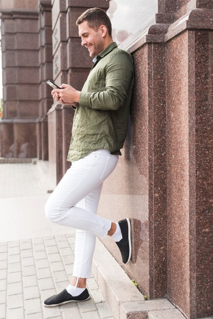 Sorrindo, homem jovem, inclinar-se parede, usando, cellphone