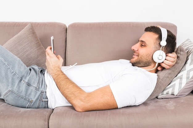 Sorrindo, homem jovem, mentir sofá, escutar música, ligado, auscultadores, através, telefone móvel