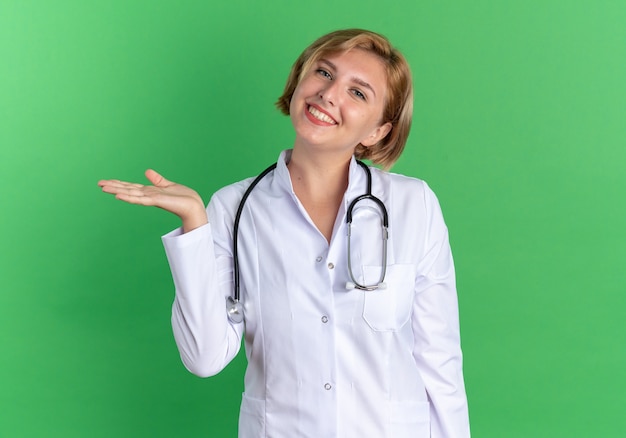 Sorrindo, inclinando a cabeça, jovem médica vestindo túnica médica com pontas de estetoscópio e a mão ao lado isolada sobre fundo verde