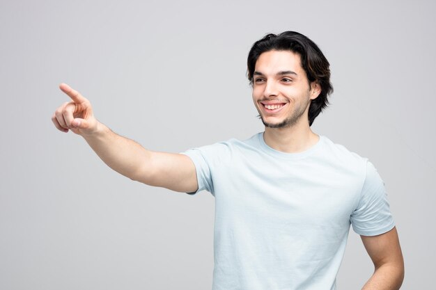 Sorrindo jovem bonito olhando e apontando para o lado isolado no fundo branco