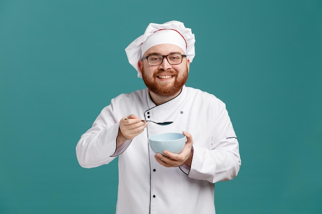 Sorrindo jovem chef masculino vestindo óculos uniforme e boné segurando tigela vazia e colher acima olhando para câmera isolada em fundo azul