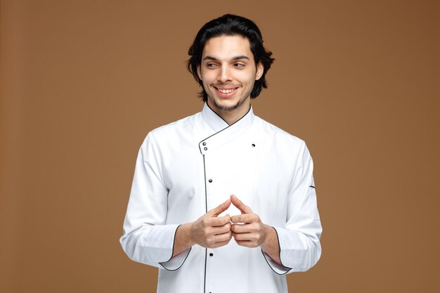 sorrindo jovem chef masculino vestindo uniforme, mantendo as mãos juntas, olhando para o lado isolado no fundo marrom
