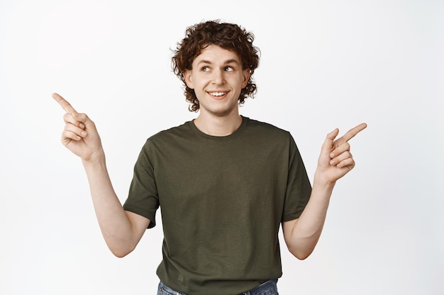 Sorrindo jovem encaracolado apontando para o lado tomando decisão escolhendo smth em pé sobre fundo branco Copie o espaço