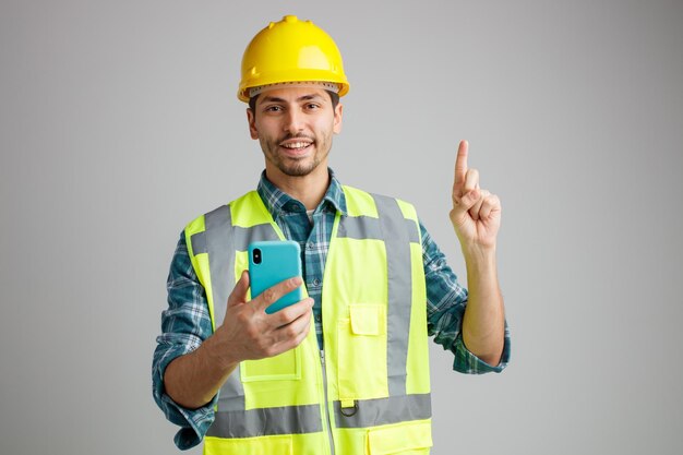 Sorrindo jovem engenheiro masculino usando capacete de segurança e uniforme segurando o celular olhando para a câmera apontando para cima isolado no fundo branco