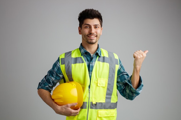 Sorrindo jovem engenheiro masculino vestindo uniforme segurando capacete de segurança olhando para a câmera apontando para o lado isolado no fundo branco