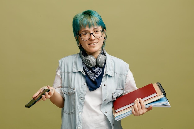 Foto grátis sorrindo jovem estudante usando óculos bandana e fones de ouvido no pescoço segurando blocos de notas e celular olhando para câmera isolada em fundo verde oliva