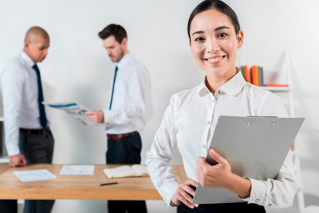 Sorrindo, jovem, executiva, segurando clipboard, em, mão, com, dois, homem negócios, trabalhando, em, fundo