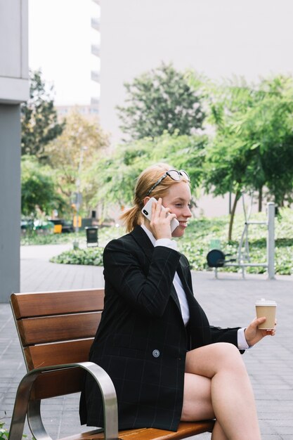 Sorrindo, jovem, executiva, sentar-se banco, segurando, taça café, takeaway, falando, ligado, telefone pilha