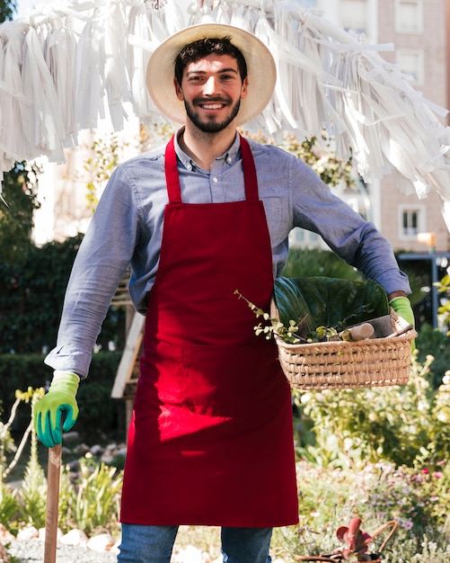 Sorrindo jovem jardineiro masculino no avental segurando cesta e ferramentas