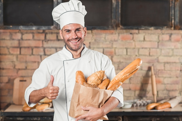 Foto grátis sorrindo, jovem, macho, padeiro, segurando, loaf, de, pães, em, saco papel, mostrando, polegar, sinal