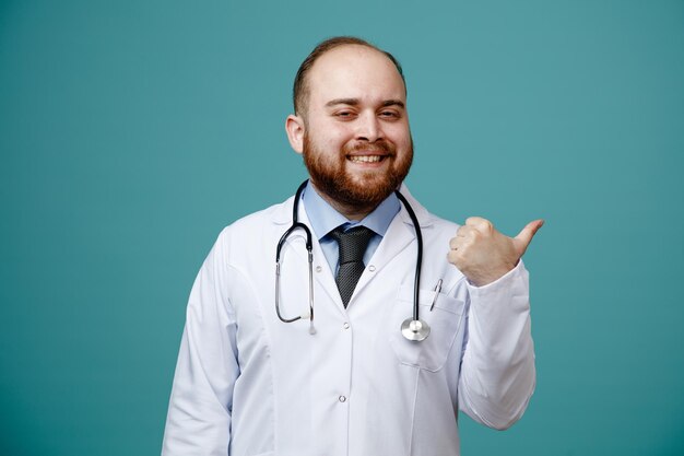 Sorrindo jovem médico masculino vestindo jaleco e estetoscópio em volta do pescoço, olhando para a câmera apontando para o lado isolado no fundo azul