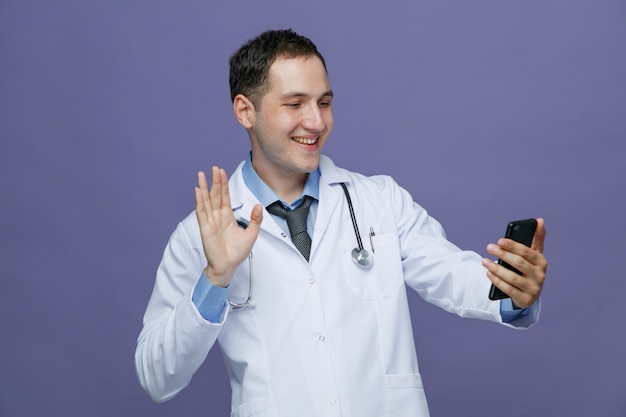 Sorrindo jovem médico masculino vestindo roupão médico e estetoscópio no pescoço esticando o celular acenando para ele falando via videochamada isolada no fundo roxo