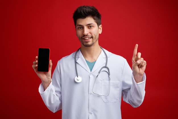 Sorrindo jovem médico masculino vestindo uniforme médico e estetoscópio em volta do pescoço, mostrando o celular para a câmera olhando para a câmera apontando para cima isolado no fundo vermelho