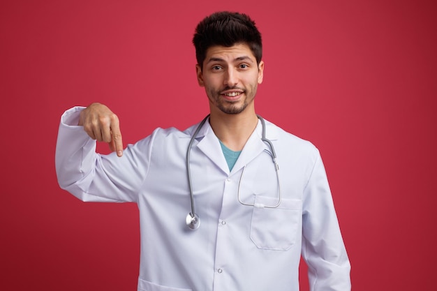 Sorrindo jovem médico masculino vestindo uniforme médico e estetoscópio em volta do pescoço, olhando para a câmera apontando para baixo