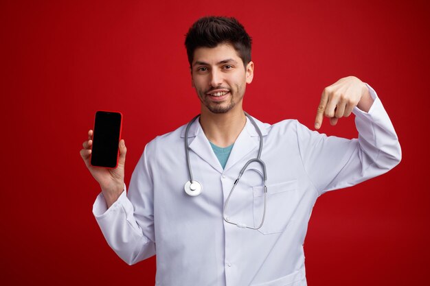 Sorrindo jovem médico masculino vestindo uniforme médico e estetoscópio em volta do pescoço, olhando para a câmera mostrando o celular apontando para baixo isolado no fundo vermelho
