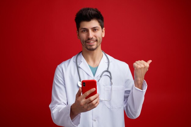 Sorrindo jovem médico masculino vestindo uniforme médico e estetoscópio em volta do pescoço segurando o celular olhando para a câmera apontando para o lado isolado no fundo vermelho