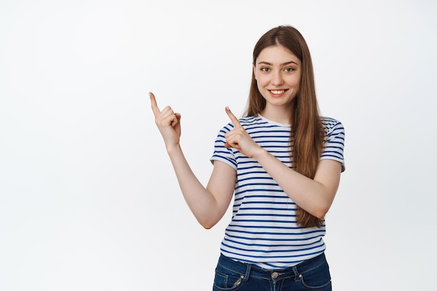 Sorrindo jovem moderna apontando para o canto superior esquerdo, mostrando propaganda, de pé em camiseta listrada contra fundo branco.