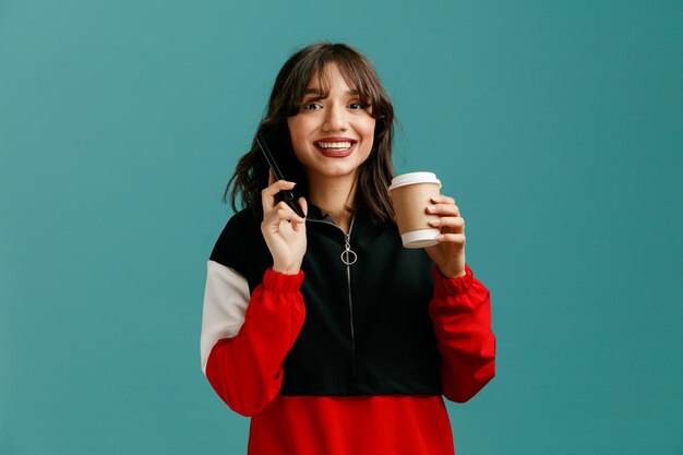 Sorrindo jovem mulher caucasiana segurando o celular perto da orelha e xícara de café para viagem em outra mão olhando para câmera isolada em fundo azul