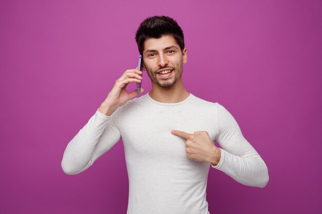 Sorrindo jovem olhando para a câmera apontando para si mesmo enquanto fala no telefone isolado no fundo roxo