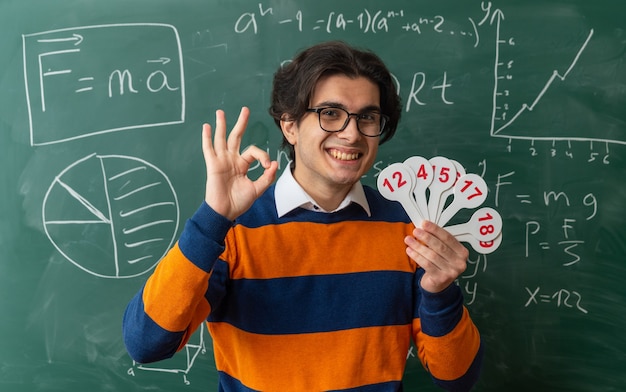 sorrindo, jovem professor de geometria usando óculos em pé na frente do quadro-negro na sala de aula segurando leques de números olhando para frente fazendo cartaz de ok