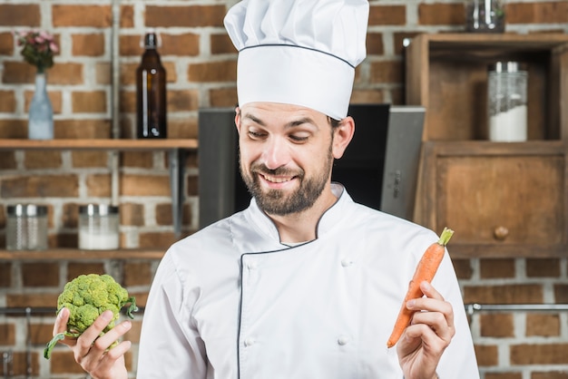 Sorrindo jovem segurando cenoura fresca e brócolis verde na cozinha