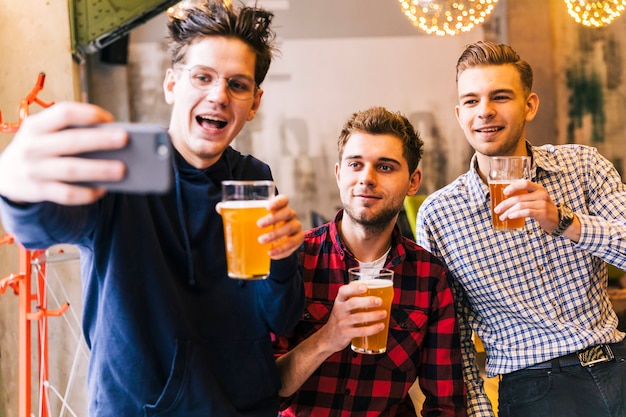 Sorrindo jovens amigos do sexo masculino segurando os copos de cerveja tomando selfie no telemóvel