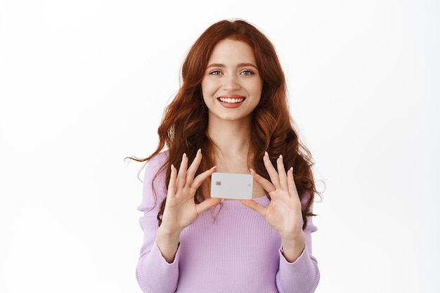 Sorrindo linda garota com cabelo ruivo mostra seu novo cartão de crédito bancário, fazendo compras em lojas sem contato, comprando coisas com dinheiro de depósito, de pé sobre fundo branco